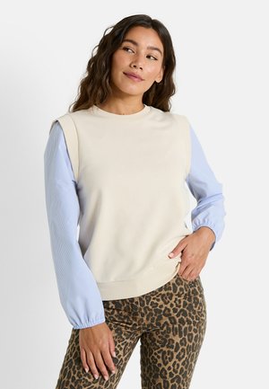 Sudadera crema con mangas a rayas azules, cuello y puños acanalados. Combinada con pantalones de estampado de leopardo, que muestran texturas y patrones contrastantes.