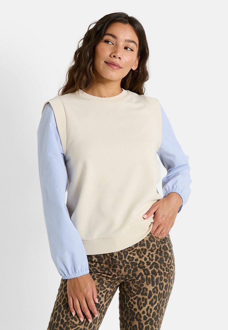 Sudadera crema con mangas a rayas azules, cuello y puños acanalados. Combinada con pantalones de estampado de leopardo, que muestran texturas y patrones contrastantes.