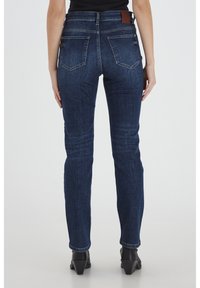 Jeans de mezclilla azul oscuro con un corte ajustado, cintura de altura media, dos bolsillos traseros y un parche de cuero marrón en la cinturilla. La textura parece suave.