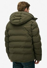Mężczyzna w zielonej, pikowanej kurtce z kapturem z logo "Superdry" na plecach, stojący tyłem na białym tle.
