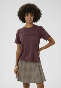 Bordeaux katoenen T-shirt met korte mouwen en de tekst "less is more" in een licht verhoogde textuur, gecombineerd met een geruite geplooide rok.