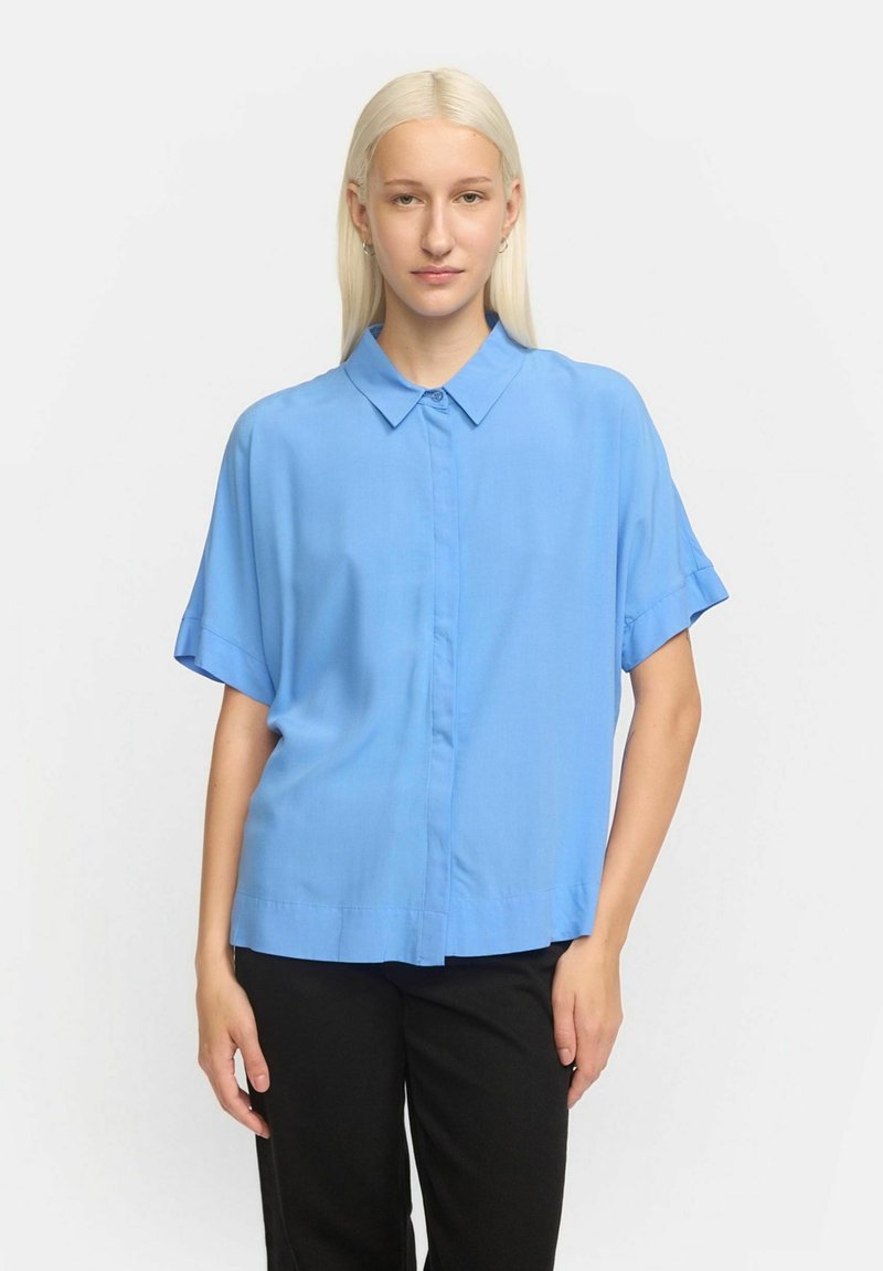 Camicia a maniche corte, con bottoni, di colore azzurro chiaro, realizzata in un tessuto liscio. Presenta un colletto strutturato e una vestibilità comoda. Indossata con pantaloni neri.