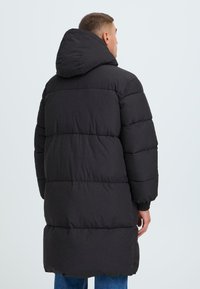 Čierna prešívaná bunda s kapucňou, dlhá, s elastickými rukávmi. Hladká textura, minimálne kovové prvky a žiadne viditeľné akcenty.