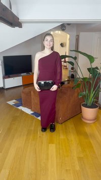 Bordó egyszáras ruha, testhezálló sziluettel és áttetsző ujjjal, fekete kézitáskával párosítva. Háttérben fa padló és zöld növény.