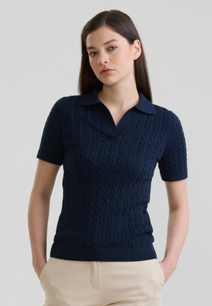 Jonge vrouw met stijl bruin haar draagt een marineblauw kabelgebreide polo met korte mouwen en beige broek, staat met handen in de zakken.