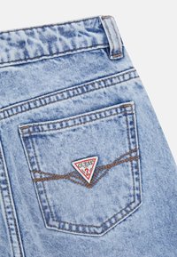Hellblaue Denim-Jeans mit brauner Naht und einem dreieckigen Guess-Markenlogo-Patch in der Mitte der Gesäßtasche.