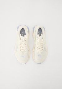 Pár smetanově zbarvených běžeckých bot Puma s světle modrými akcenty, viděný shora na bílém pozadí.