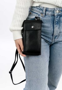 Black leather crossbody phone bag with a front snap pocket, adjustable strap, and textured finish, held in a hand against light denim.