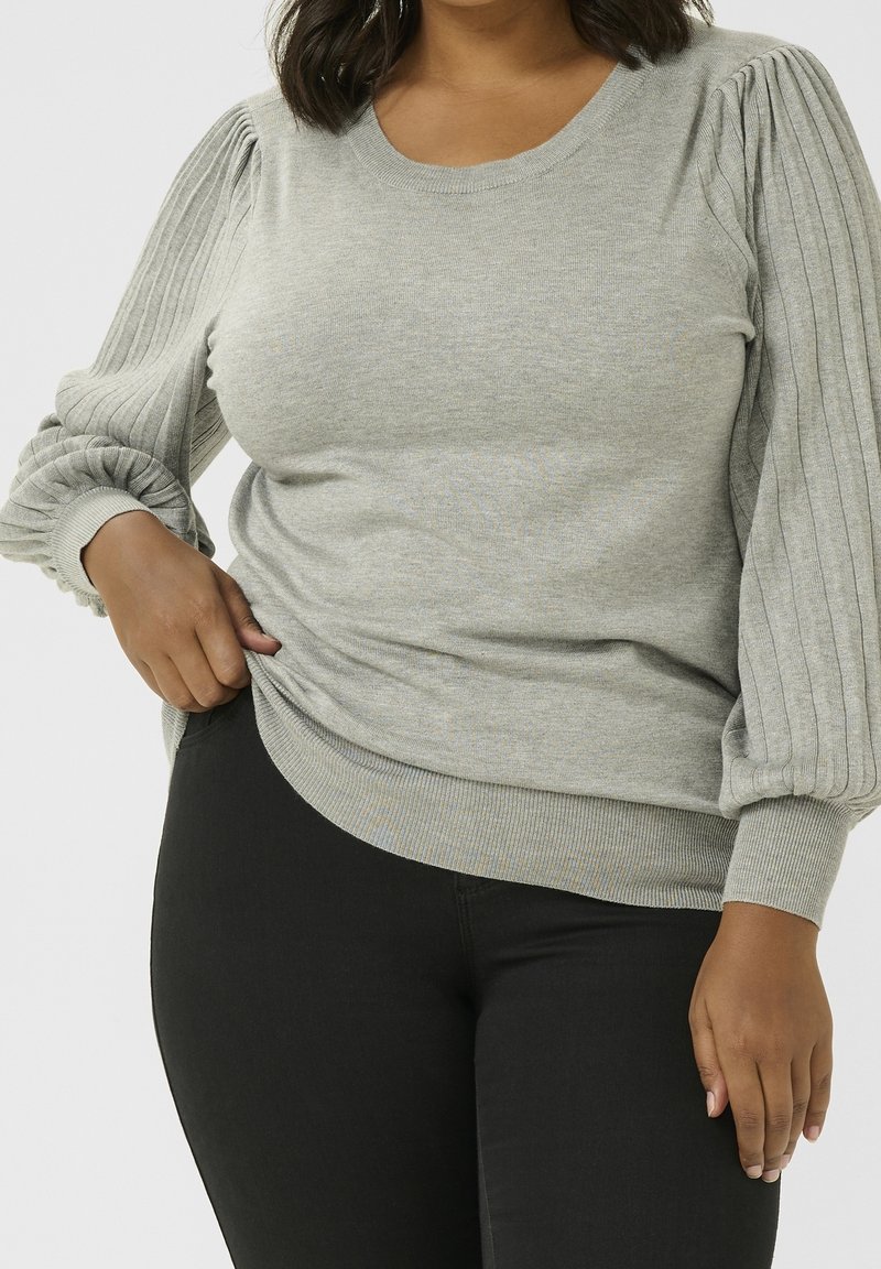 Femme portant un pull gris clair côtelé à manches longues bouffantes et un pantalon noir, tenant l'ourlet du pull de la main gauche.