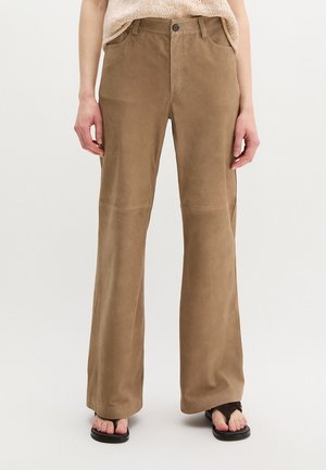 Vrouw die bruine wijde broek met hoge taille en voorzakken draagt, gecombineerd met zwarte teenslippers, tegen een witte achtergrond.