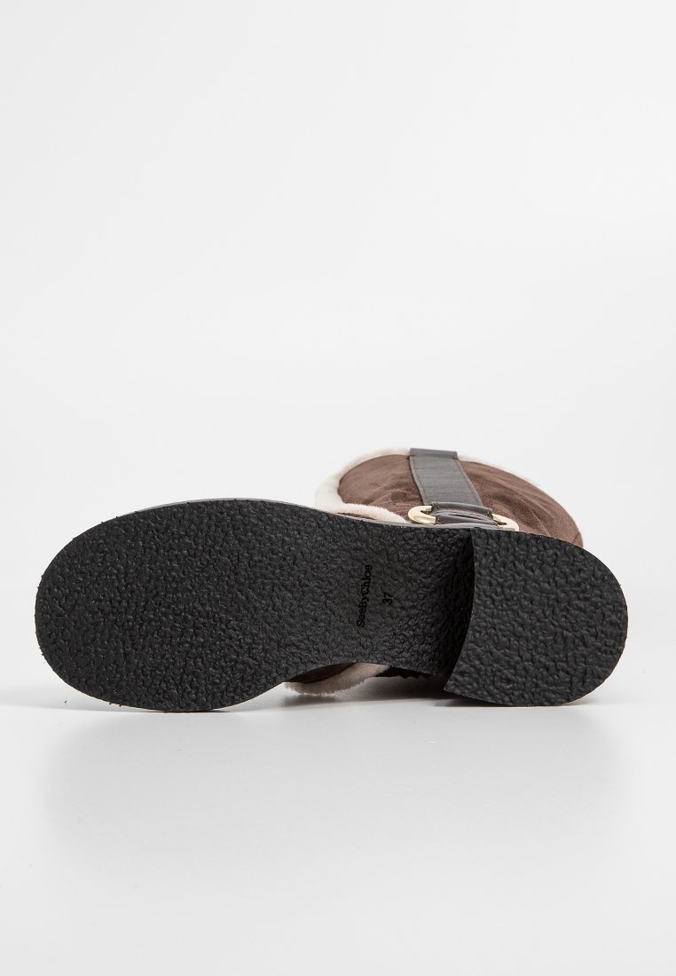 Bruine suede sandaal met een gestructureerde zwarte rubberen zool, verstelbare band en zachte bontvoering. Bevat een cirkelvormige gouden accent nabij de gesp.