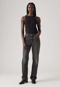 Zwarte geribbelde tanktop en high-waisted donkergrijze jeans met rechte pijpen. Model draagt zwarte haklaarzen. Soepele textuur, minimale details.