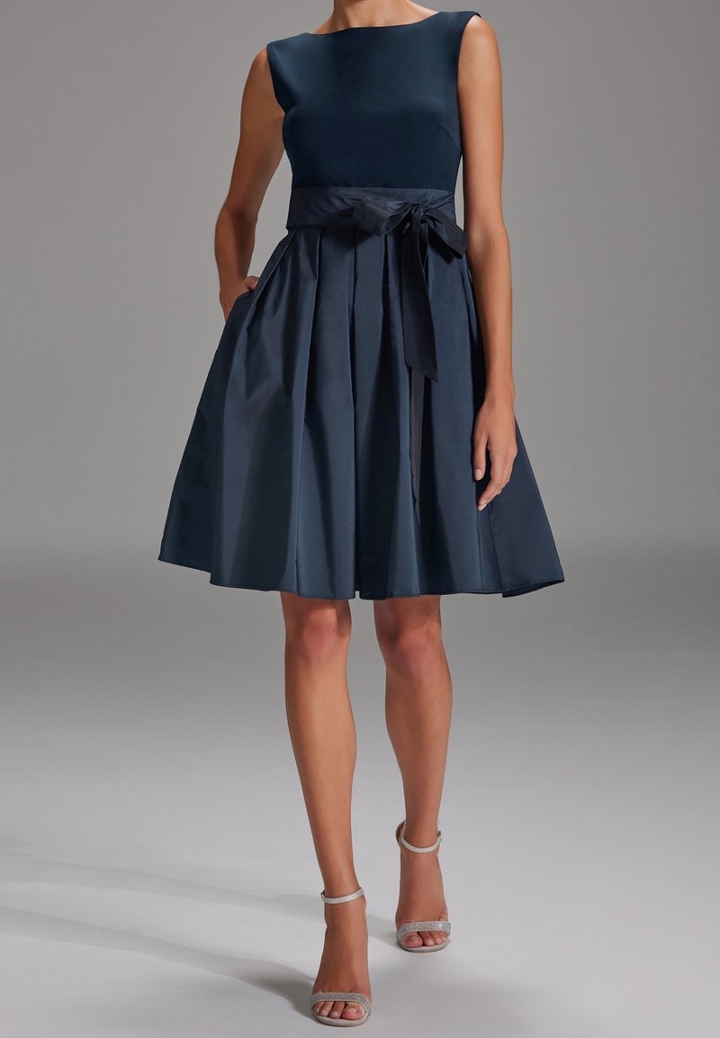Robe sans manches bleu marine avec un corsage ajusté, une jupe plissée et un ruban noué à la taille. Tissu lisse avec une finition brillante.