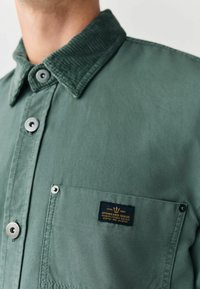 Grüne Hemd mit einem Cordkragen, Metallknöpfen und einer Brusttasche, die ein marineblaues Etikett mit goldener Schrift besitzt. Glatte Textur und taillierte Passform.