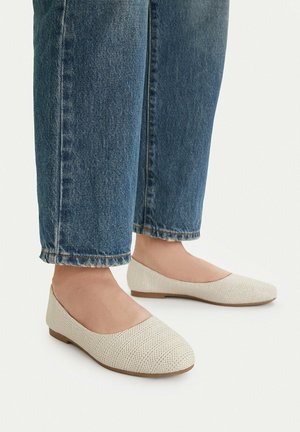 Stopy w białych, teksturowanych balerinach zestawionych z niebieskimi dżinsami o prostych nogawkach na jasnym tle.