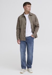 Veste button-up marron clair avec un col pointu, portée sur un t-shirt blanc, associée à un jean bleu et des baskets de couleur claire.