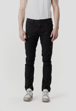 Pantalones cargo negros de corte ajustado, con múltiples bolsillos, detalles con cremallera y una textura suave. Combinados con zapatillas blancas y grises.