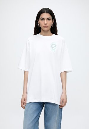 Γυναίκα με μακριά σκούρα μαλλιά φοράει μια μεγάλη λευκή μπλούζα με μικρό πράσινο σχέδιο και ανοιχτό μπλε τζιν, στέκεται μπροστά σε μια απλή λευκή επιφάνεια.