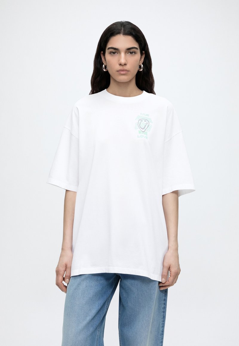 Γυναίκα με μακριά σκούρα μαλλιά φοράει μια μεγάλη λευκή μπλούζα με μικρό πράσινο σχέδιο και ανοιχτό μπλε τζιν, στέκεται μπροστά σε μια απλή λευκή επιφάνεια.