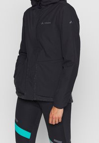 Svart vattentät jacka med en strukturerad inre foder, med huva, framfickor och en logotyp på bröstet; strukturerad tyg och stilren design.