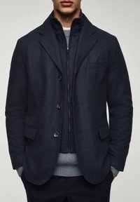 Man in een donkerblauwe blazer over een gesloten marineblauwe jack en een lichtgrijze trui, met de handen in de zakken, getoond van schouders tot heupen.