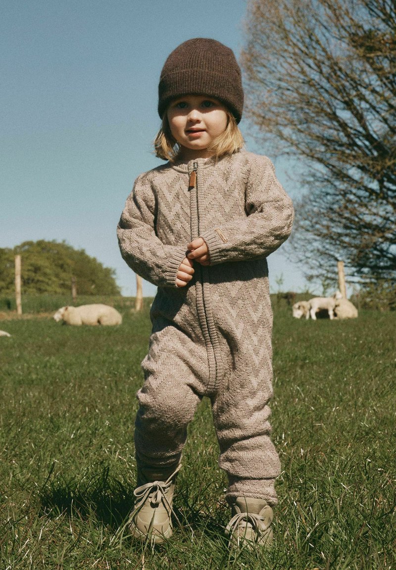 Jeune enfant portant un bonnet marron en tricot et une combinaison beige, debout sur l'herbe avec des moutons et des arbres en arrière-plan sous un ciel dégagé.