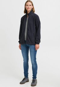 Marineblå fleecejakke med høy krage, glidelås foran og to sidelommer, kombinert med grå t-skjorte, blå jeans og grå joggesko.
