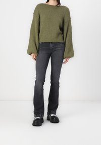Pull en tricot surdimensionné vert olive avec des manches larges, associé à un jean gris foncé  à pattes d'eph et des chaussures noires à semelles épaisses.