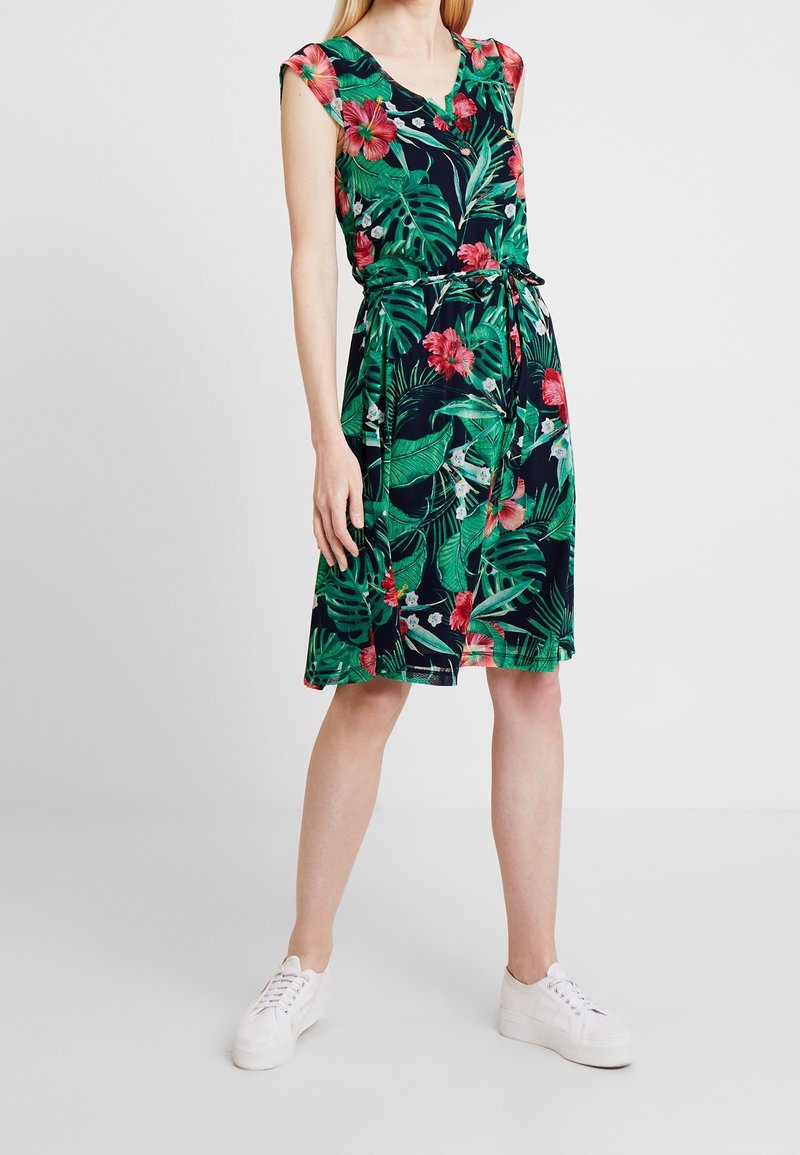 Robe sans manches jusqu'aux genoux avec imprimé feuilles tropicales vertes et fleurs d'hibiscus roses, ceinture nouée à la taille, portée avec des baskets blanches.