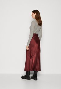 Fustă maxi din satin burgund cu o textură netedă, cu talie înaltă și tiv asimetric, purtată cu un top cu dungi cu mâneci lungi și cizme negre.
