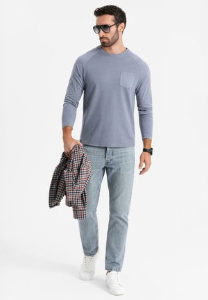 Lichtblauwe blouse met lange mouwen en een borstzak, gecombineerd met lichtblauwe jeans en witte sneakers. Een geruite flanellen blouse is over de arm gedrapeerd.