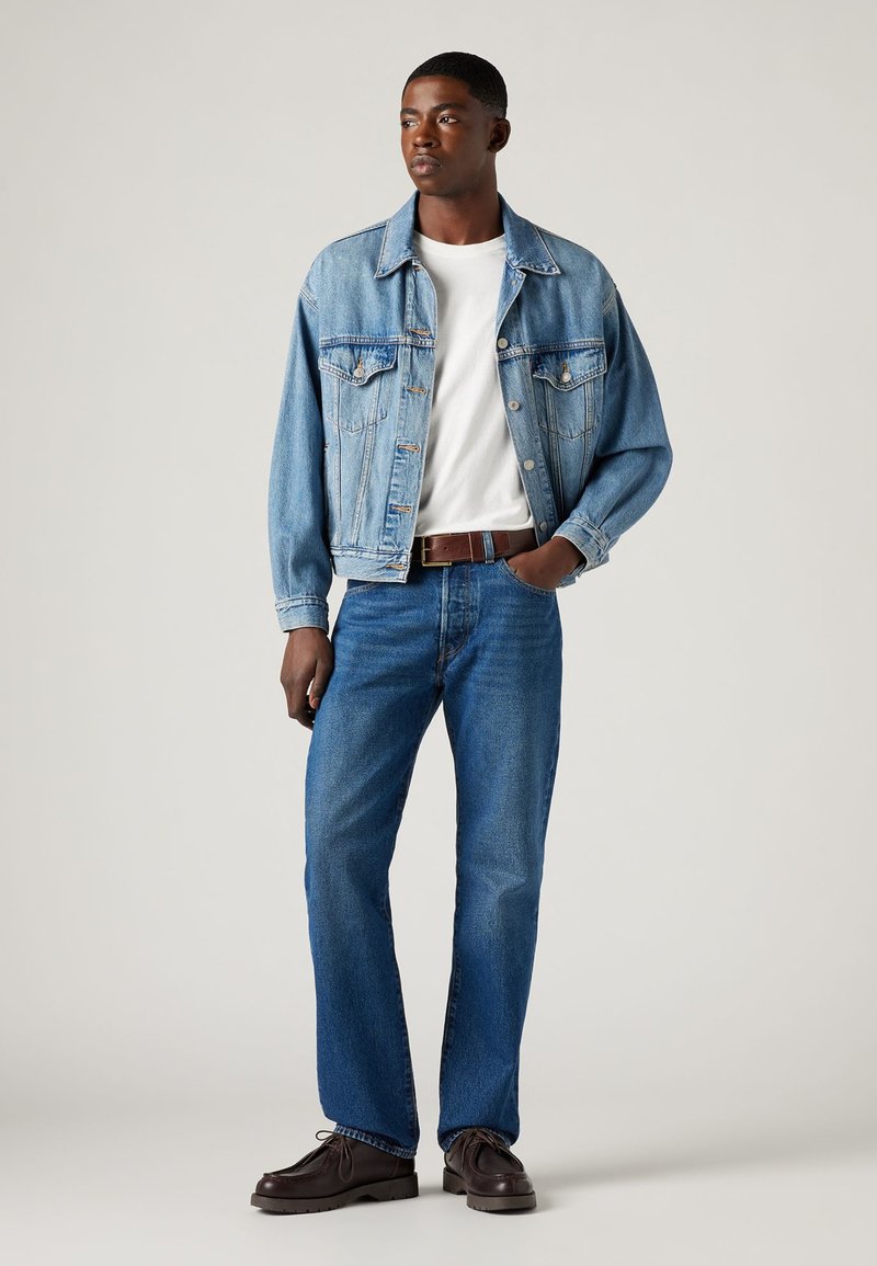 Hellblaues Jeansjackett über einem weißen Hemd, kombiniert mit blauen Jeans und braunen Schnürschuhen. Einfaches Design mit klassischen Hardware-Details.
