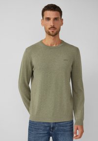 Olivgrüner Strickpullover mit Rundhalsausschnitt, langen Ärmeln und gerippten Bündchen. Mit einem kleinen Logo auf der Brust. Kombiniert mit blauen Jeans.