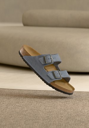 Sandale en daim bleu avec deux boucles argentées, semelle intérieure en liège et semelle noire flottant au-dessus d'un tapis beige et d'un canapé assorti.