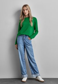 Grön stickad tröja, vida ljusblå denimjeans med uppvikta ben, vita sneakers. Modellen poserar avslappnat mot en grå vägg.