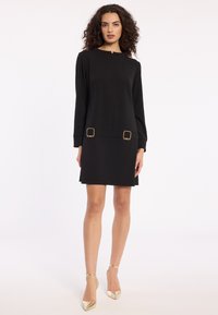 Woman standing in a black long-sleeve mini dress with gold buckle details at the waist, wearing gold pointed-toe heels.