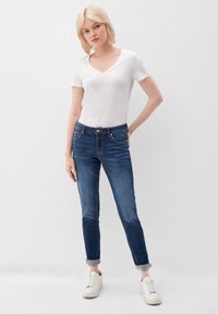 Camiseta blanca de escote en V y jeans ajustados azul oscuro con un ligero desgaste, puños enrollados y bolsillos frontales, combinados con zapatillas blancas.