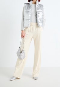 Silberne gepolsterte Weste über einem grauen Oberteil, kombiniert mit cremefarbenen weit geschnittenen Hosen. Die Accessoires umfassen silberne Spitzenschuhe und eine schimmernde Handtasche.