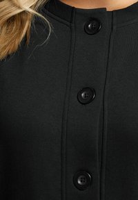 Prenda negra de botonadura frontal con una textura suave, un escote redondo y cuatro grandes botones negros a lo largo de la abertura.