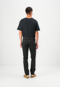 Zwarte jeans en een donker T-shirt, gezien van achteren. De jeans hebben twee achterzakken; het model draagt bruine schoenen met witte accenten.