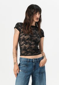 Haut en dentelle noire à manches courtes avec une silhouette ajustée, présentant un motif ondulé et un col rond. Associé à un jean en denim bleu clair.