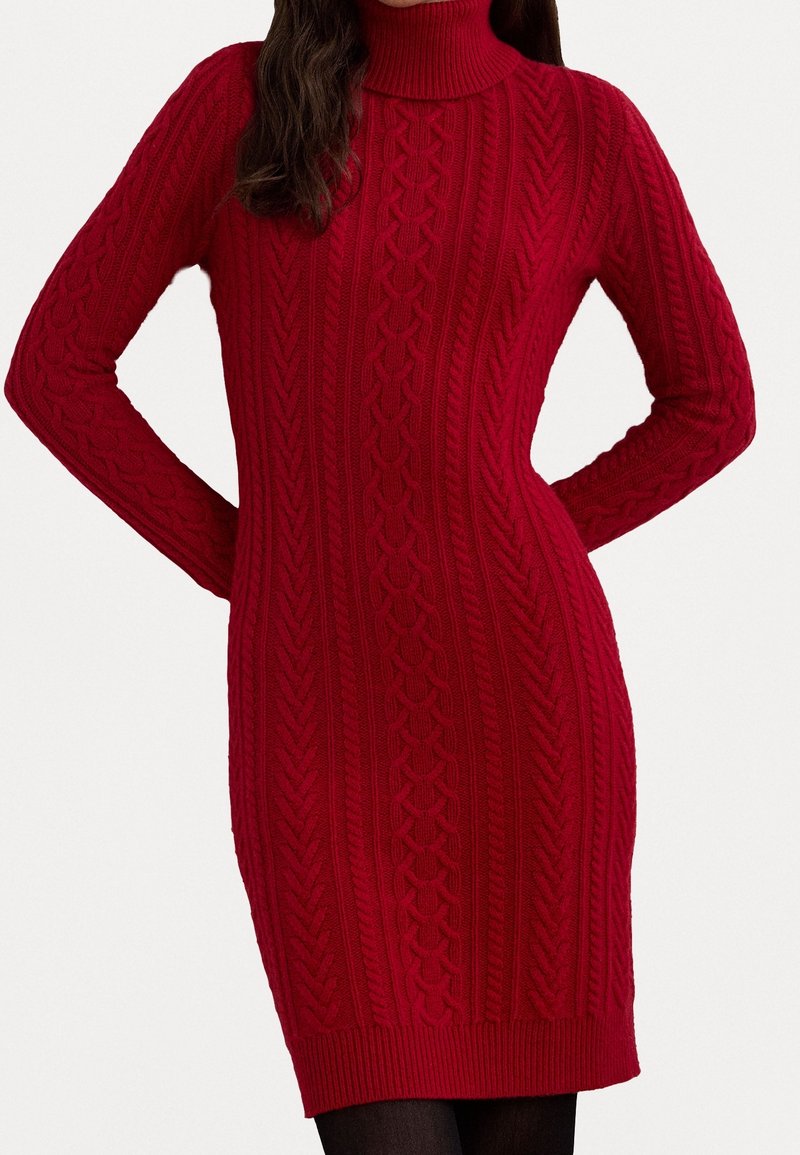 Robe pull à col roulé rouge en maille torsadée avec manches longues et motifs verticaux texturés, portée avec des collants noirs.