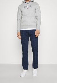 Šedá mikina s logem "TOMMY HILFIGER", bílá košile pod ní, námořnické kostkované kalhoty a bílé tenisky. Neformální outfit s volným střihem.