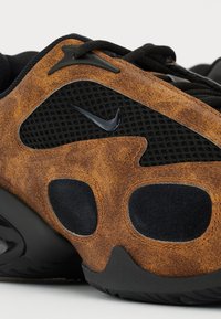 Gros plan sur une sneaker Nike marron et noire, montrant le tissu en maille, les panneaux en cuir texturé et le logo swoosh noir sur le côté.
