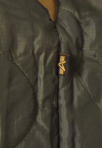 Gros plan sur un tissu de veste matelassée olive foncé avec une petite étiquette de marque tissée noire et jaune cousue dans la couture.