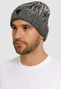 Cappello a maglia grigio con motivi a onde nere, texture a coste nella parte inferiore e dettaglio logo triangolare sulla parte anteriore.