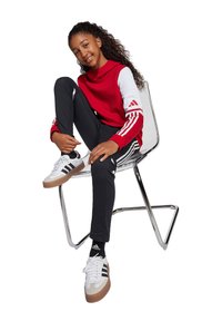 Rode en witte hoodie met drie strepen op de mouwen, zwarte broek en witte sneakers met zwarte strepen, zittend op een chrome stoel.