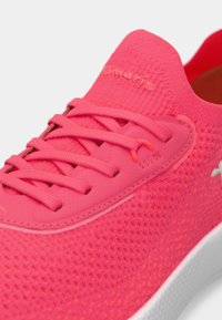 Φωτεινό ροζ αθλητικό παπούτσι με υφασμάτινο πάνω μέρος με υφή, επίπεδες κορδόνια και μαξιλαρωμένη σόλα. Αξιοσημείωτη σήμανση είναι παρούσα στο πλάι.