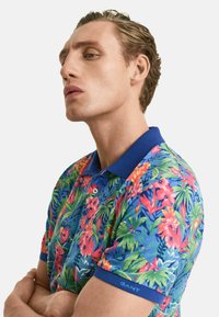 Buntes Poloshirt mit kurzen Ärmeln und blauem Kragen, das ein lebendiges Blumenmuster in Rot-, Pink-, Grün- und Blautönen zeigt. Akzent-Logo auf dem Ärmel.