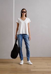 Weißes V-Ausschnitt-T-Shirt, blaue Jeans, weiße Sneakers und eine schwarze geflochtene Tragetasche. Die Umgebung hat eine hellgraue Wand und einen Holzboden.
